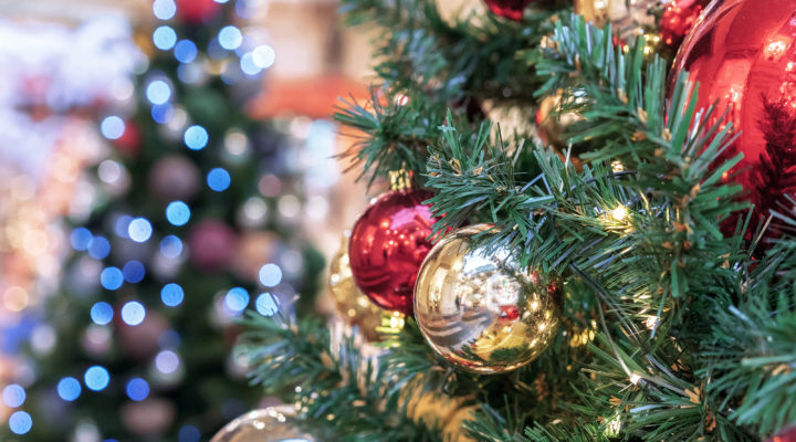 red and gold ornaments hang on a Christmas tree against the backdrop of a lighted Christmas tree bokeh